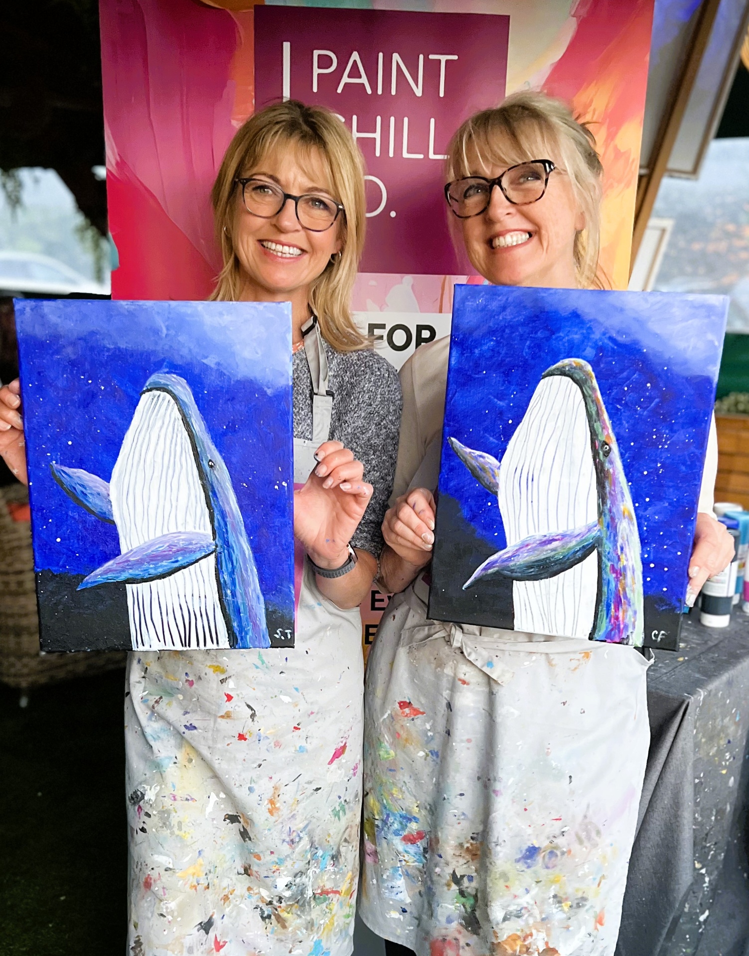 Two customers smiling with their Blue Whale paintings at a paint & sip event